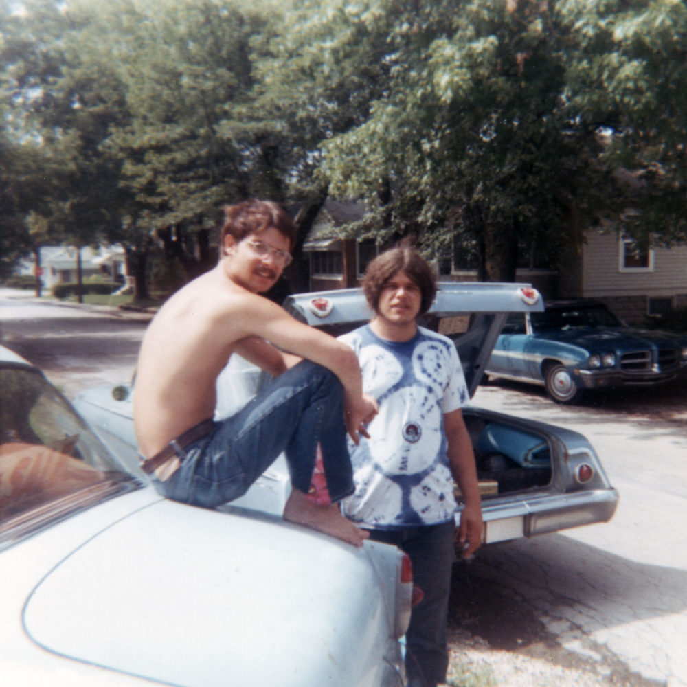 Norm sitting on his car with Brad in front of trailer on Monon St. 